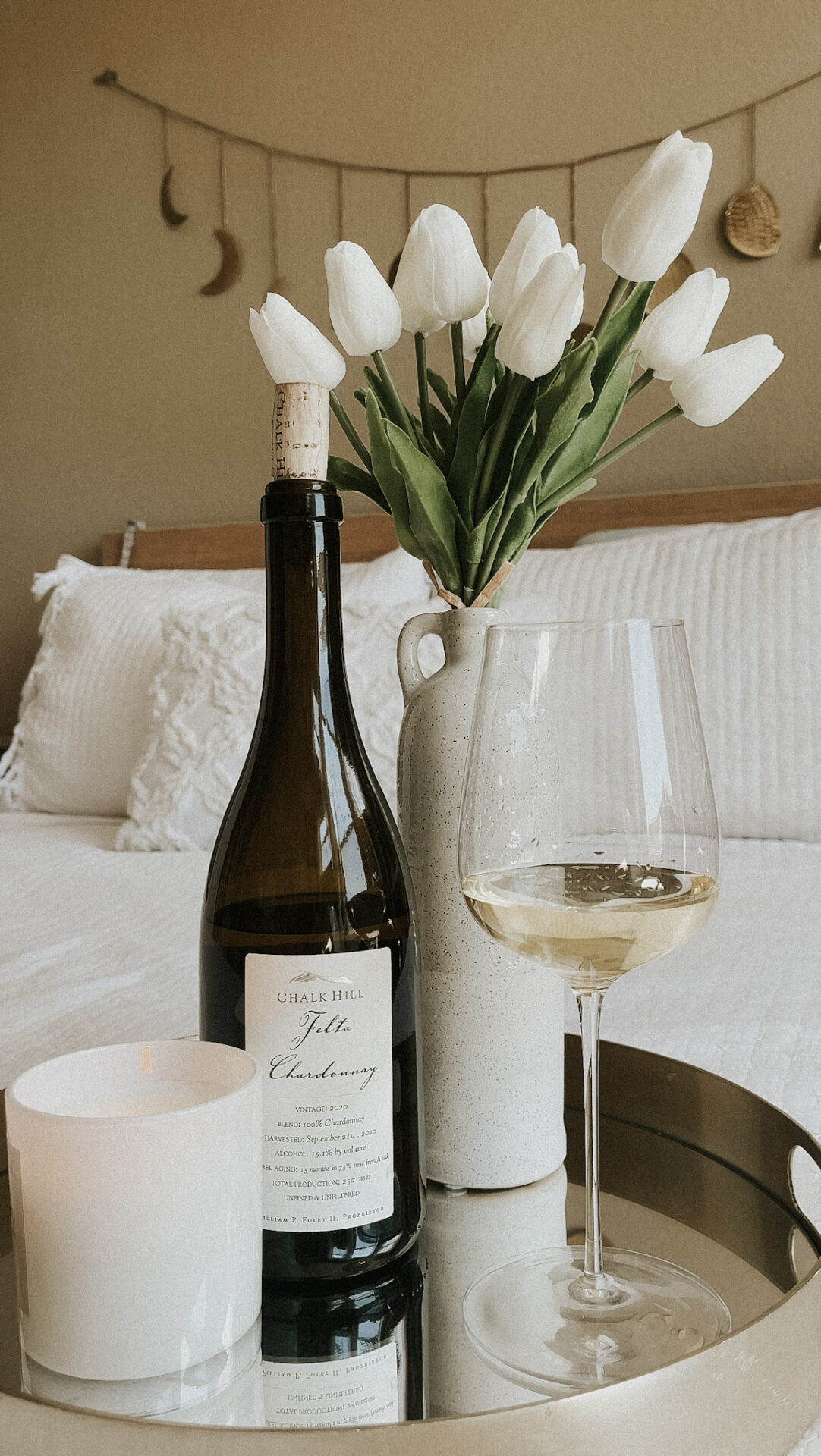 A bottle of Chardonnay on a try with a glass of wine next to it, a candle, and a vase of white tulips.