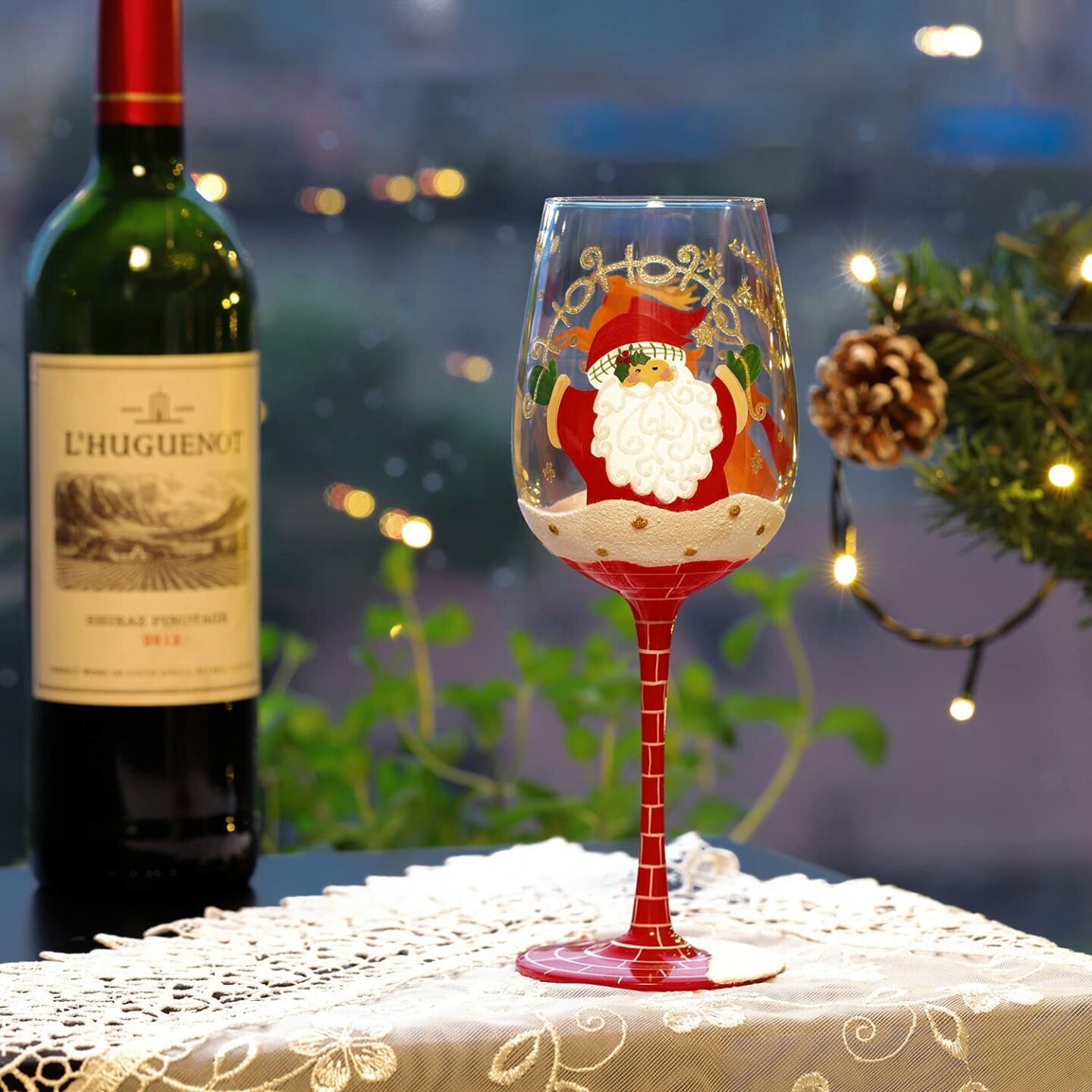 Painted wine glass of Santa coming down the chimney, pictured in front of a bottle of wine and a Christmas tree