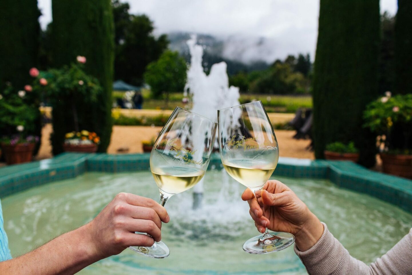 Two hands cheer wine glasses in front of a bubbling fountain