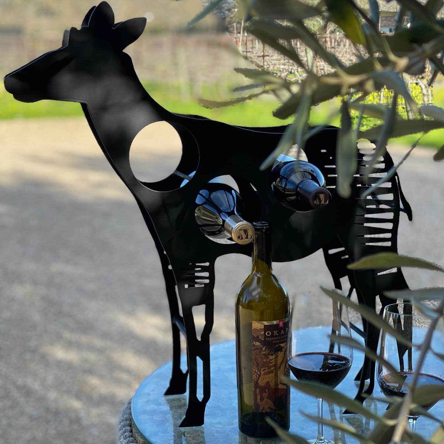 A wine holder, in the shape of an okapi, holds two bottle of wine, and another bottle is photographed on the table beside it