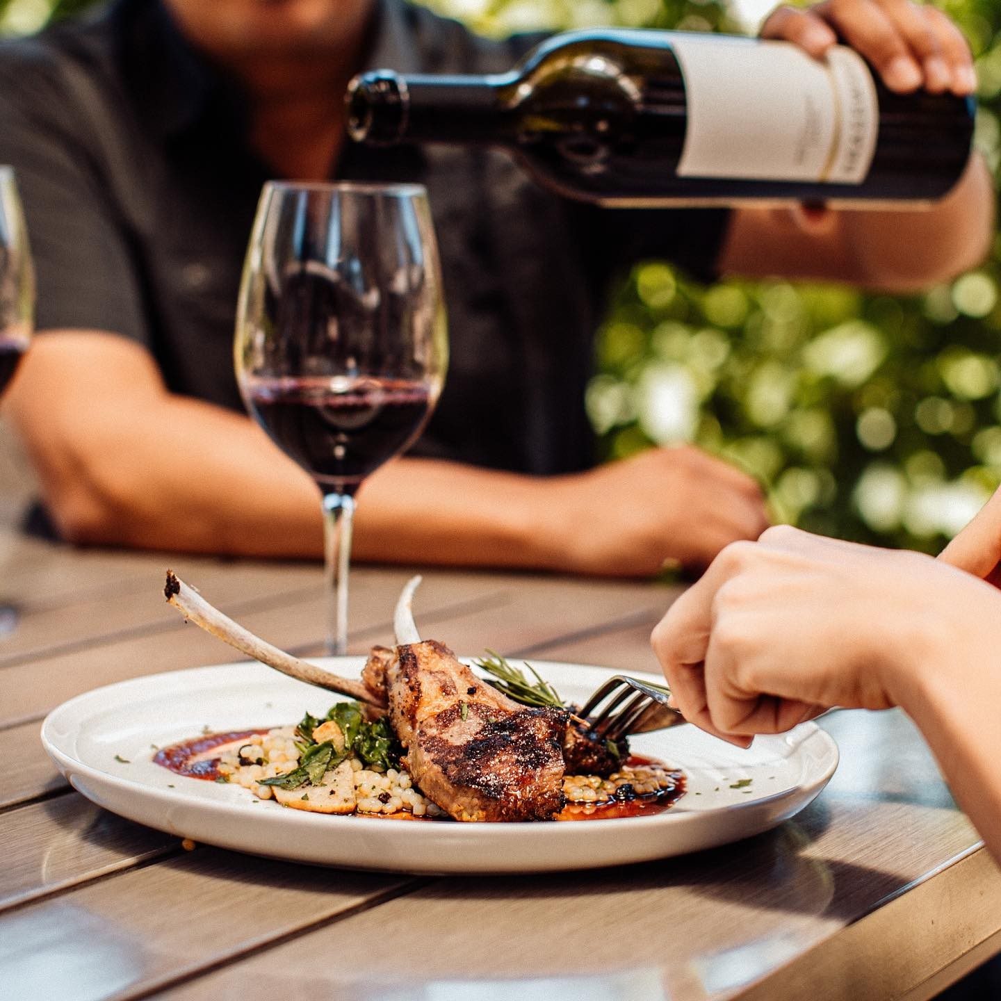 A hand cutting into a pork chop while another pours a glass of red wine to pair at Brasswood