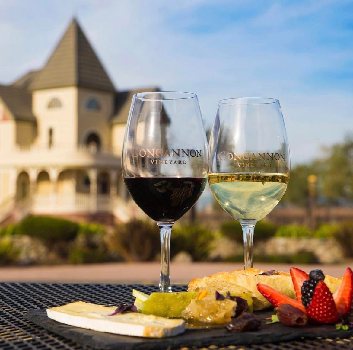 Two glasses of wine and a plate of fruit photographed with a large Victorian home in the background at Concannon Vineyard in Livermore, CA