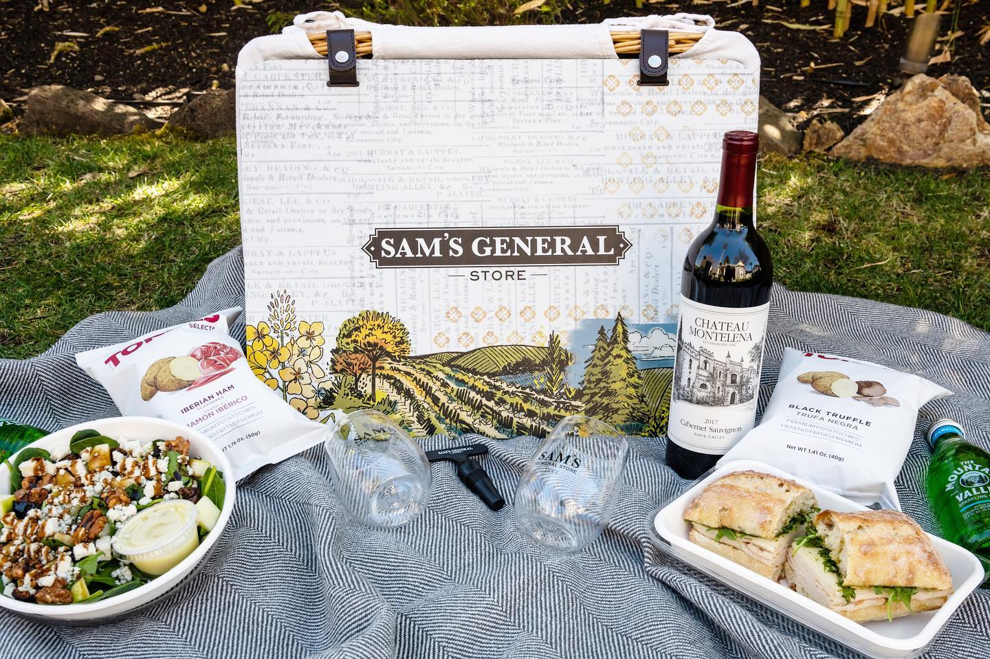 A picnic basket featuring sandwiches, salads, cookies and a bottle of wine set up on a blanket outside