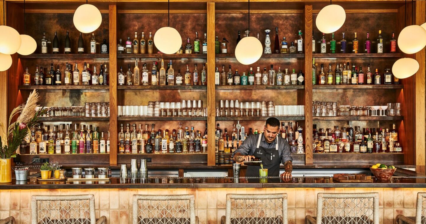 A bartender pours a drink standing behind the counter of a fully stocked bar