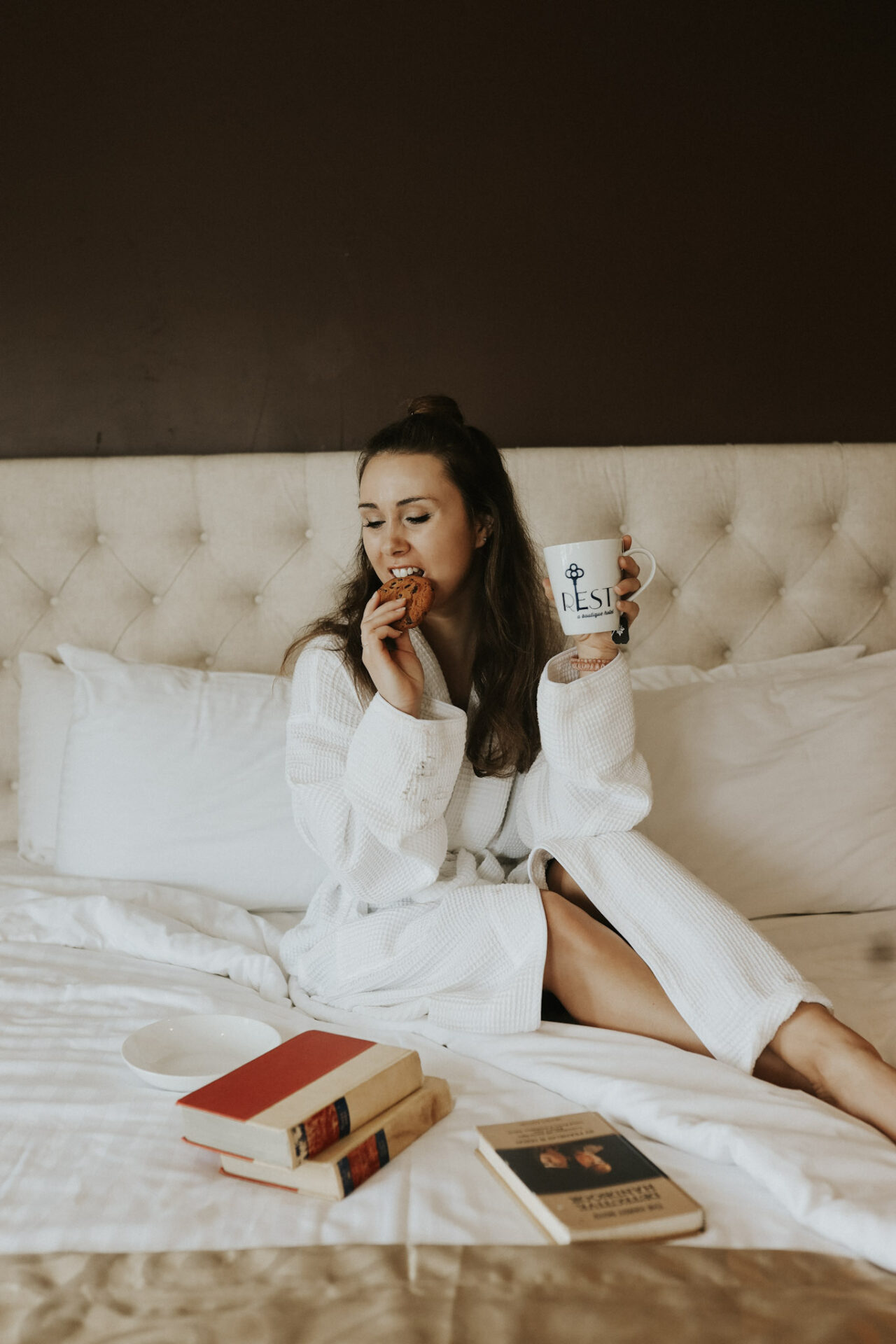 Paige on a bed at Rest Hotel in Amador County. Coffee Cup in one hand with a cookie in another and vintage books on the bed