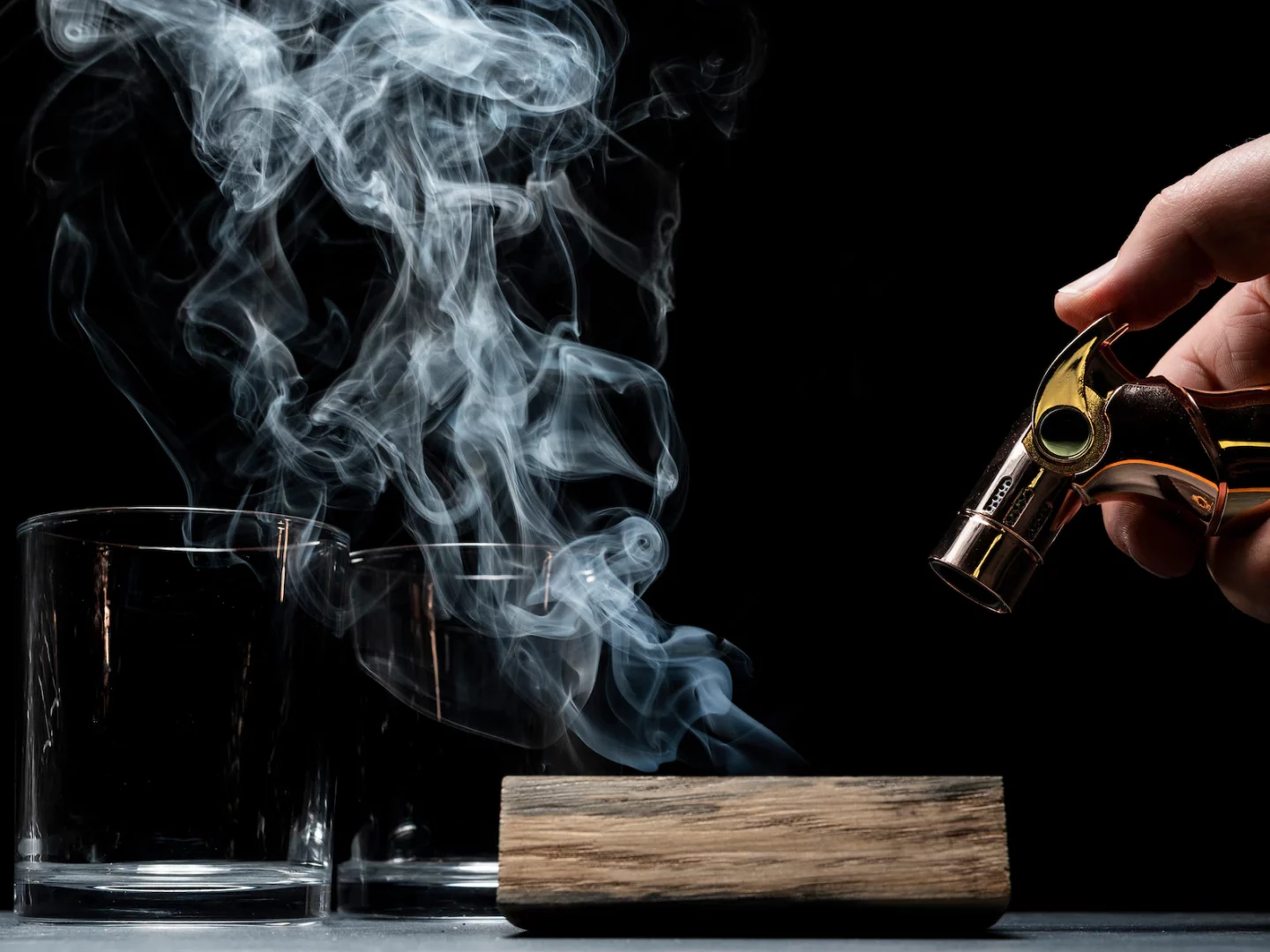 A hand holds a culinary blow torch over a smoking slab of wood, set beside two cocktail glasses