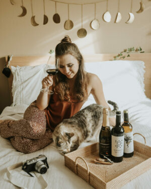 Paige drinking a GSM Wine blend with three bottles of wine in front of her on a bed and her cat, Arwen. A camera is shown also on the bed.