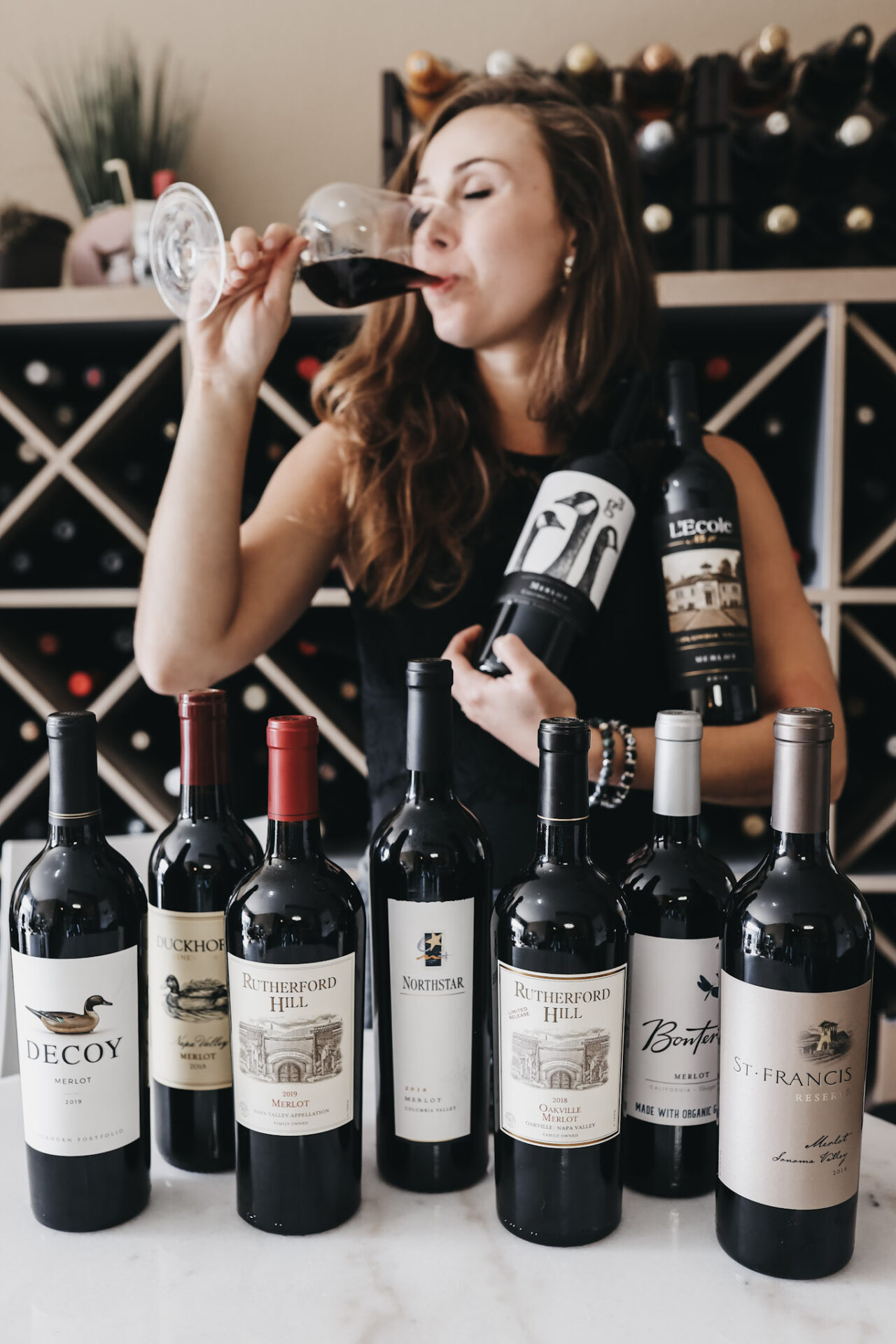 Paige surrounded by Merlot bottles, drinking red wine