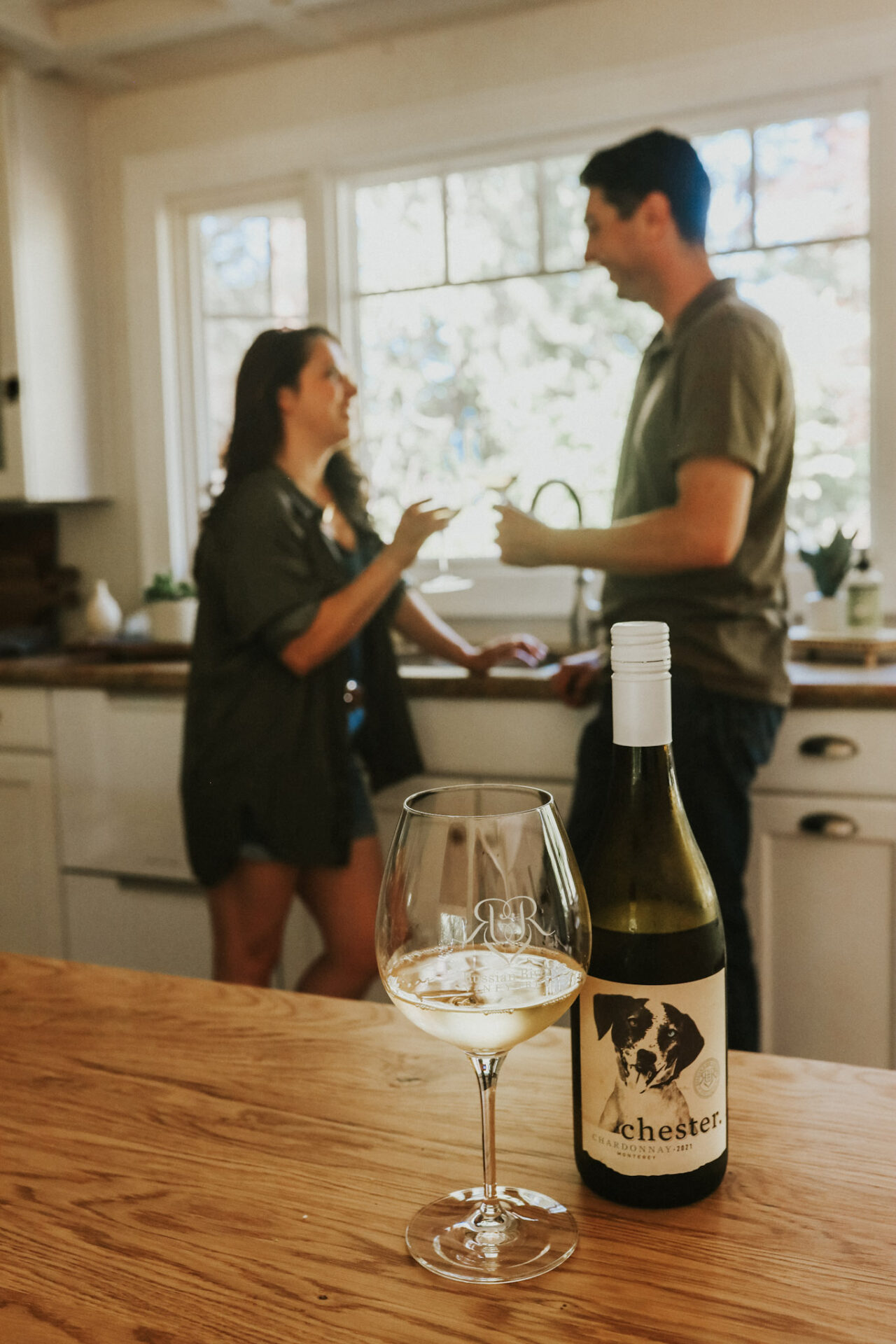 A glass of unoaked Chardonnay next to a bottle on a kitchen counter. A couple stands in the background