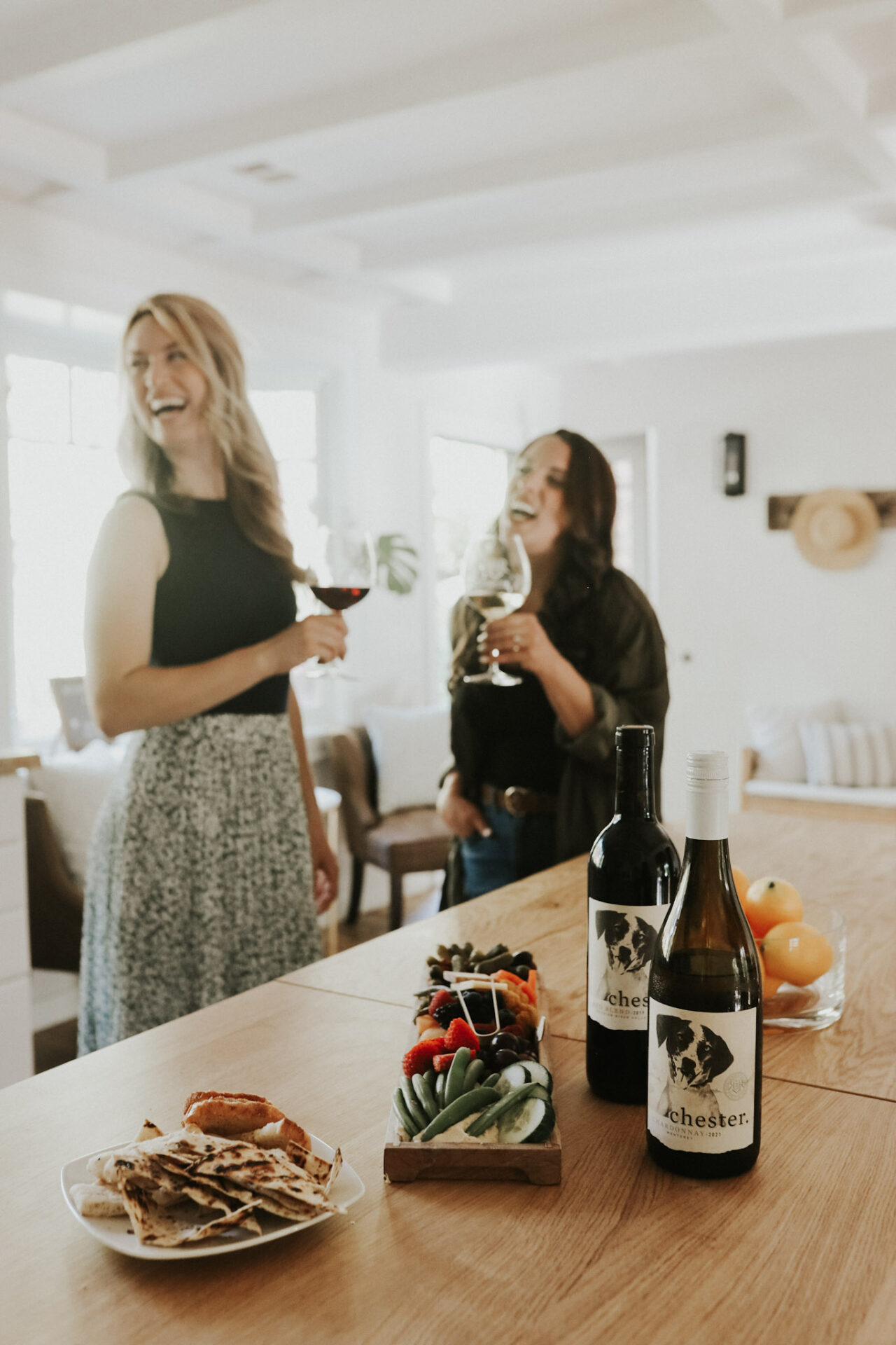 two friends drinking wine with charcuterie in the foreground and two bottles of wine, including chardonnay.