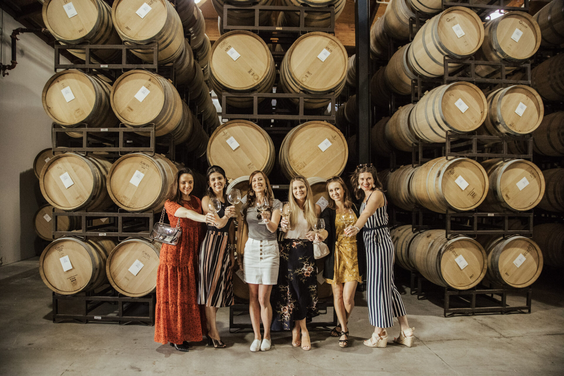 A group of women with oaked chardonnay in wine glasses in front of wine barrels